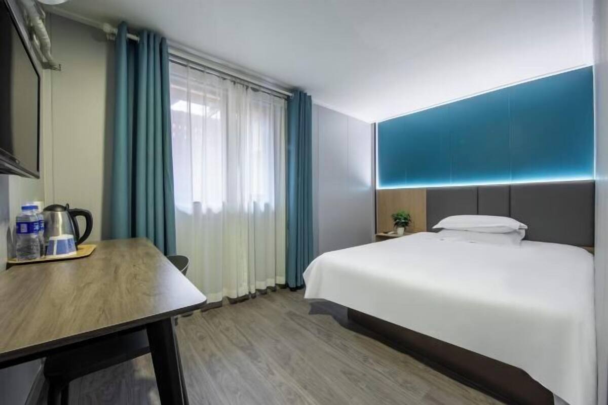Une chambre d’hôtel avec un lit, un bureau avec une bouilloire, une petite table avec des bouteilles d’eau et une table de chevet avec une plante.