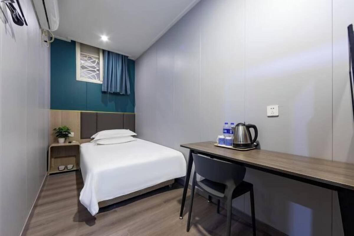 Une chambre d’hôtel comprenant un lit, un bureau, une chaise, une petite table avec une bouilloire, une plante et une prise électrique.