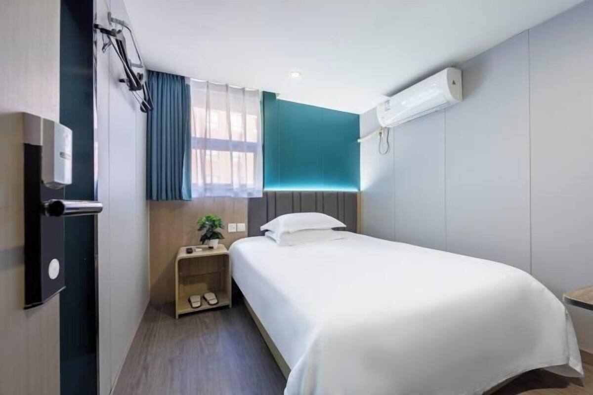 Une chambre d’hôtel avec un lit, une petite table de chevet avec des chaussons, un climatiseur fixé au mur et une fenêtre avec des rideaux.