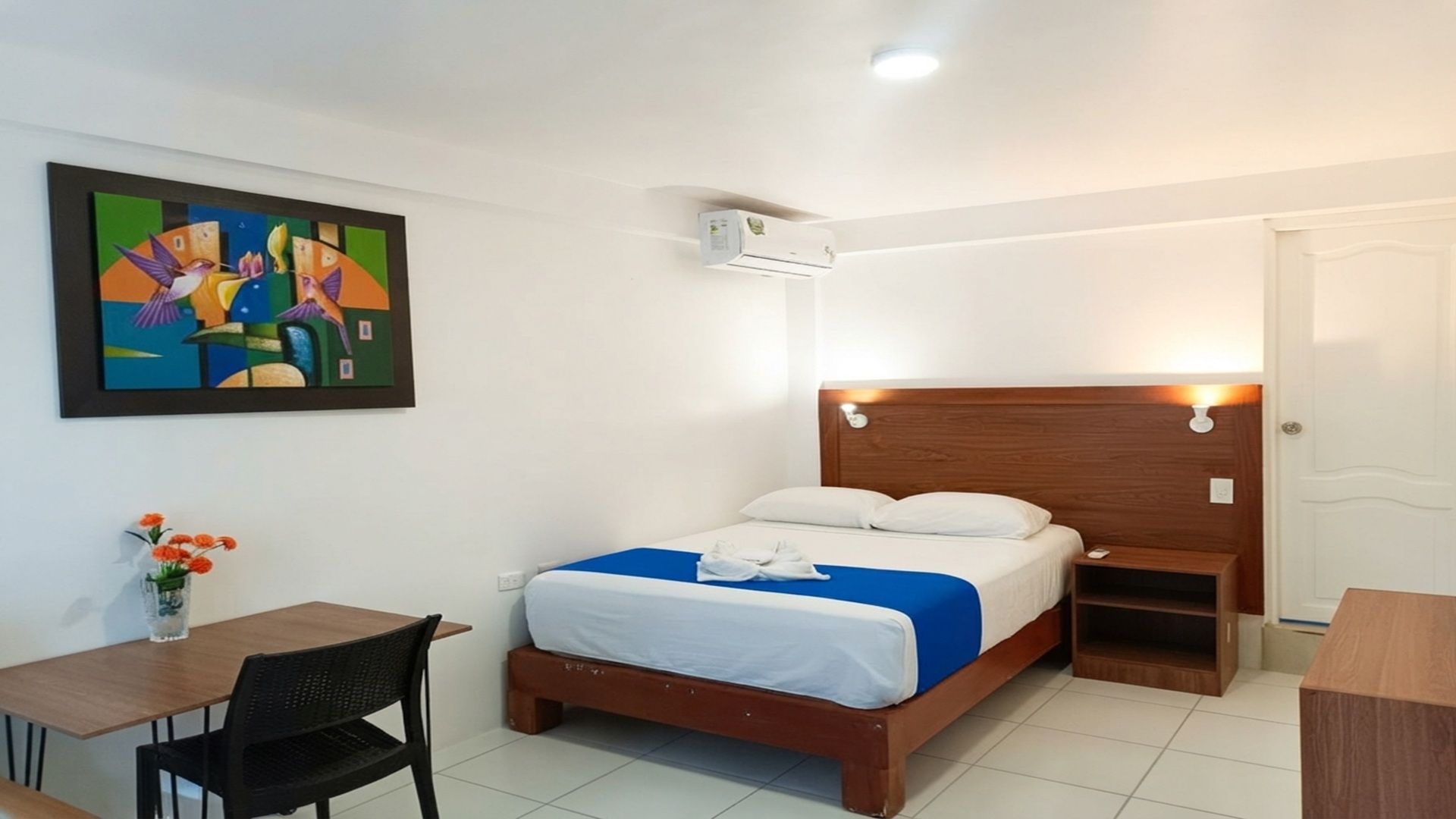 A hotel room with a bed, bedside table, chair, and a painting on the wall.