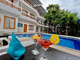 Zona de piscina con dos bebidas sobre una mesa, una piscina y un edificio con varios balcones.