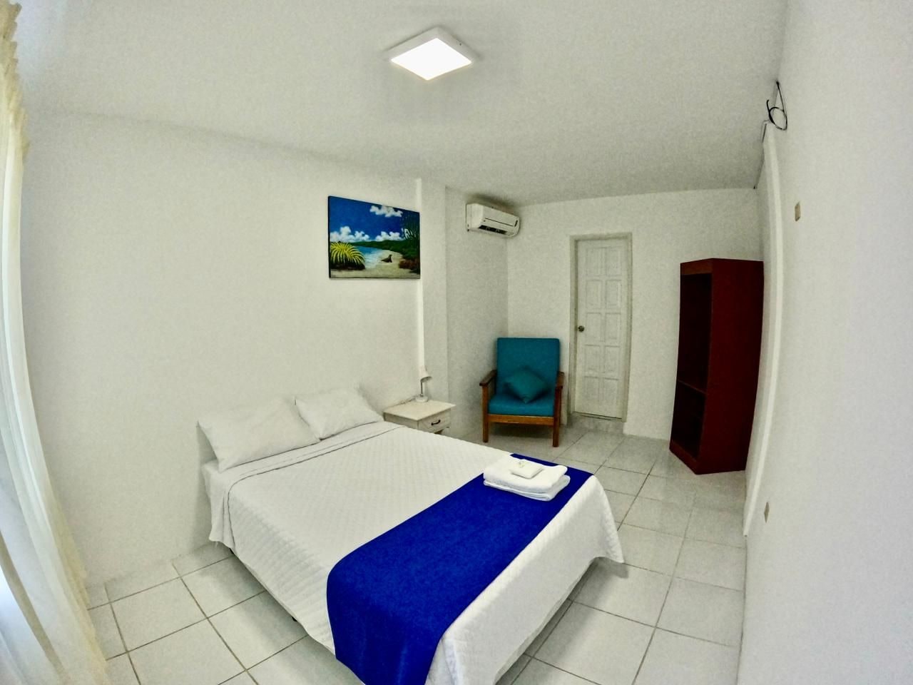 A hotel room with a bed, a chair, a wardrobe, and an air conditioning unit.