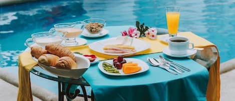 A breakfast setup by a pool with croissants, fruit, coffee, and juice.