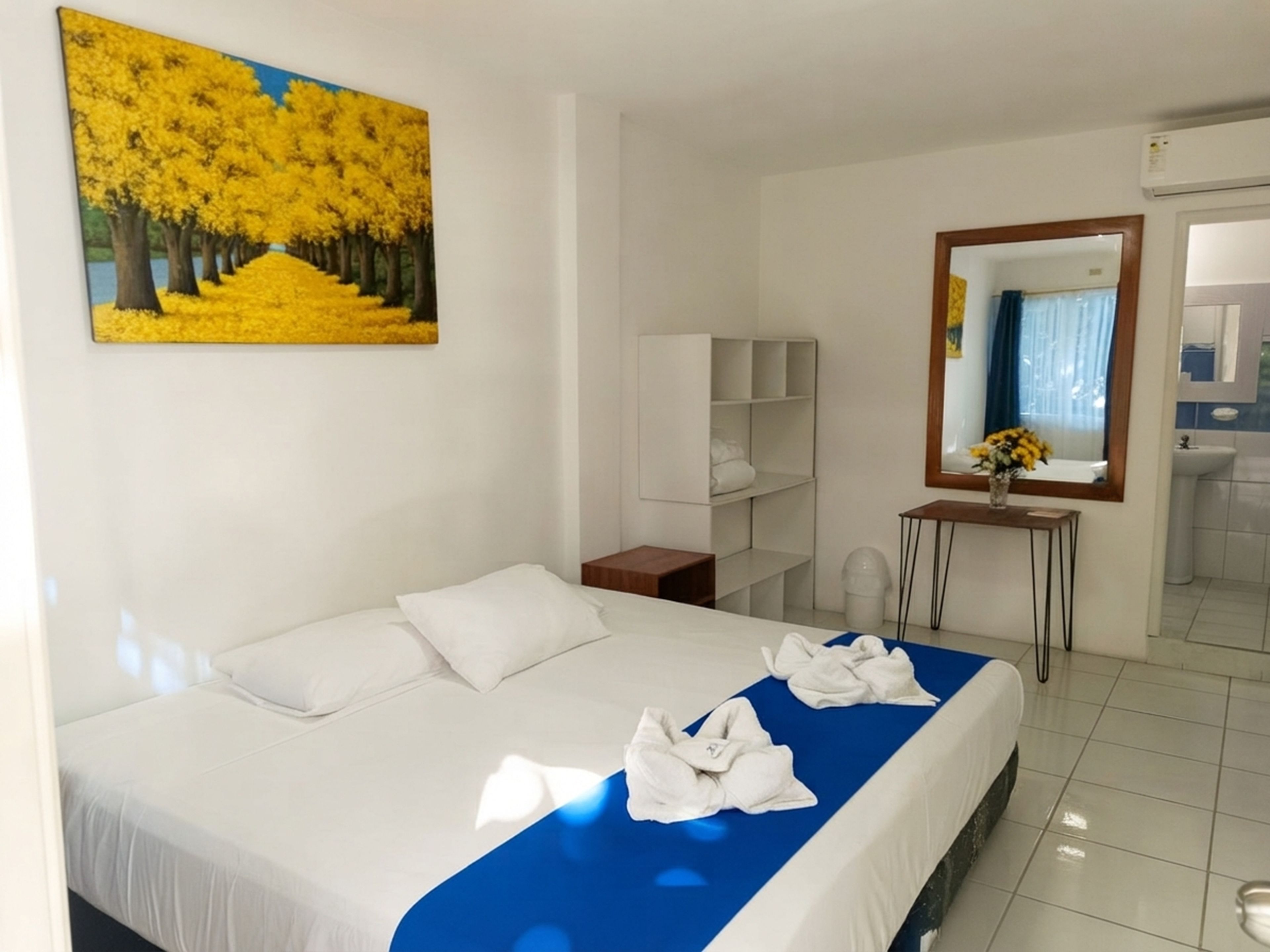 A hotel room with a bed, a painting, a mirror, a small table with a vase, and a bathroom visible through an open door.