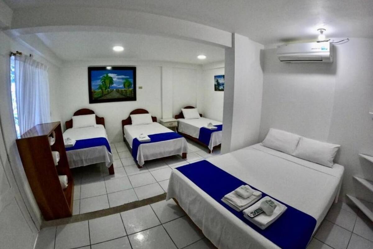 A hotel room with two single beds and one double bed, white bedding, a blue accent on the beds, a painting on the wall, and an air conditioning unit.