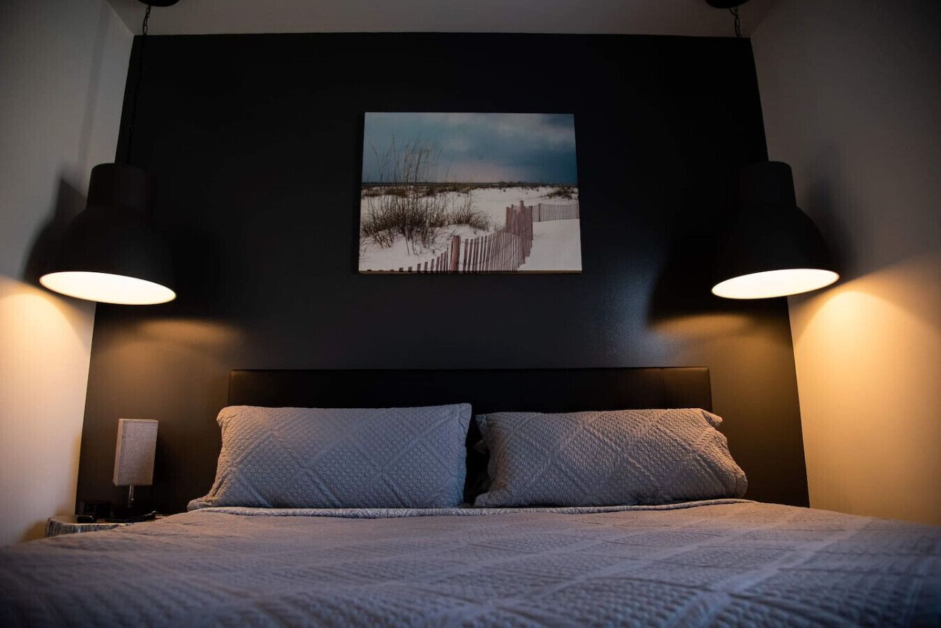Een bed met twee kussens, een donker hoofdbord en erboven een lijst met een strandafbeelding.
