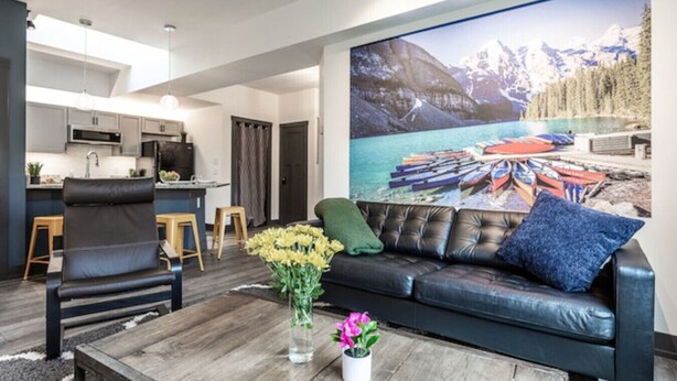 A modern living room with a black leather sofa, a wooden coffee table, and a large painting of a lakeside scene. - Grand Park Modern Cubby 2BR/3BED/1BA/patio (Westfield)