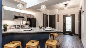 A modern kitchen with granite countertops, stainless steel appliances, and pendant lighting. There are three stools at the island. - Grand Park Modern Cubby 2BR/3BED/1BA/patio (Westfield)