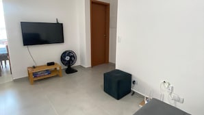 Un salon doté d’un téléviseur à écran plat fixé au mur, d’un meuble de rangement en bois, d’un ventilateur noir et d’un pouf de couleur sombre.