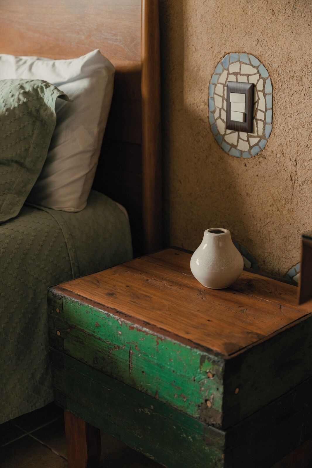 A wooden nightstand with a white vase, a green and brown chest, and a mosaic wall decoration.