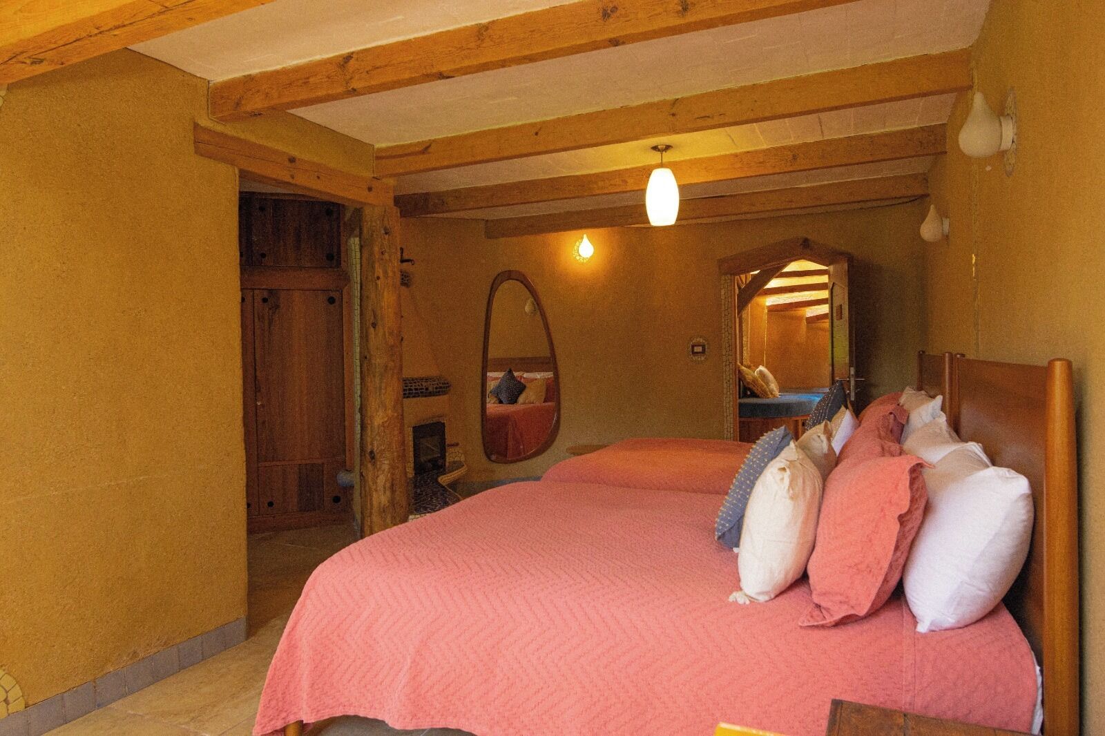 Une chambre confortable avec un plafond en bois, un grand lit avec une literie rose, un fauteuil suspendu et un miroir au mur.