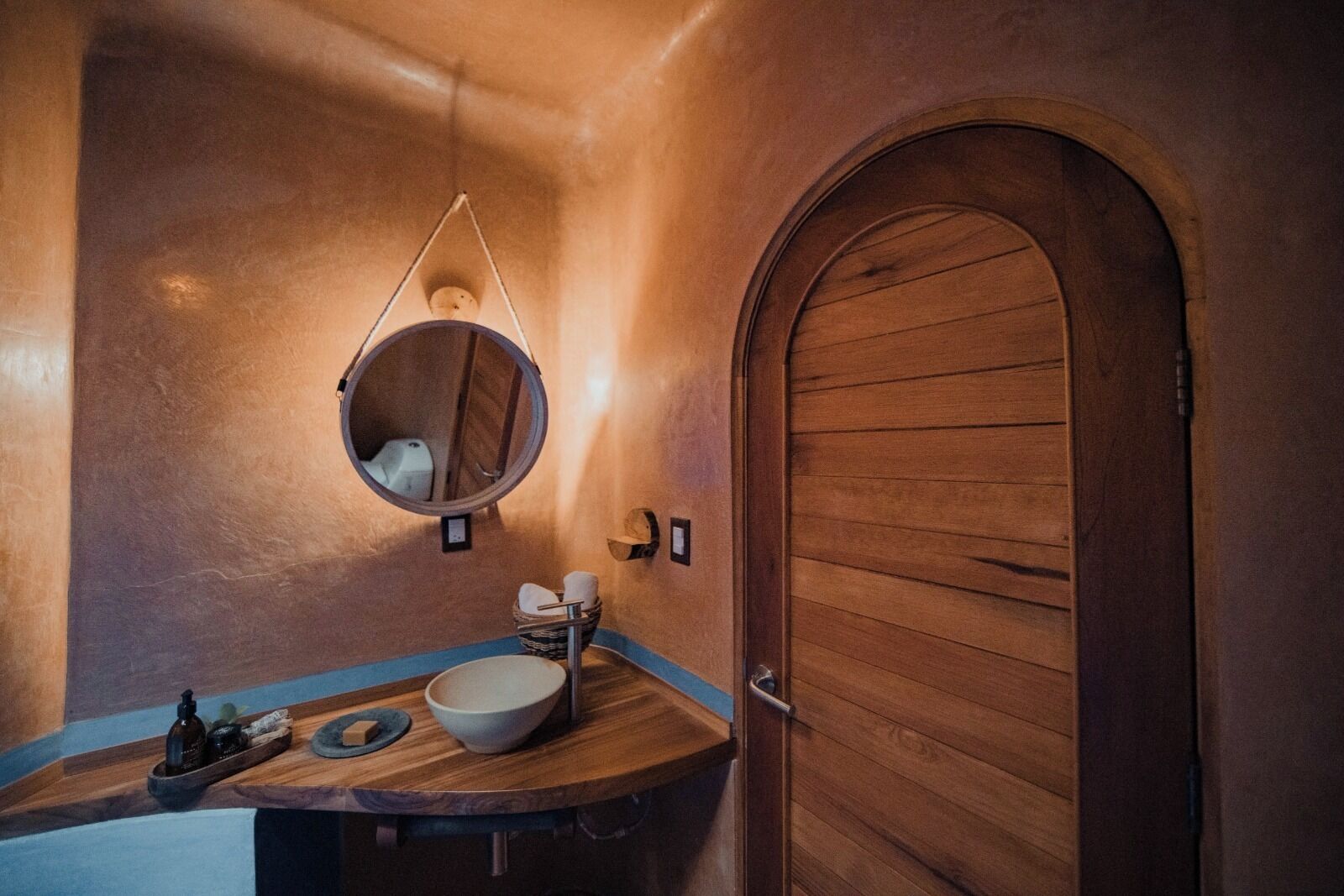 A bathroom with a wooden door, a round mirror, a sink, and a towel rack.