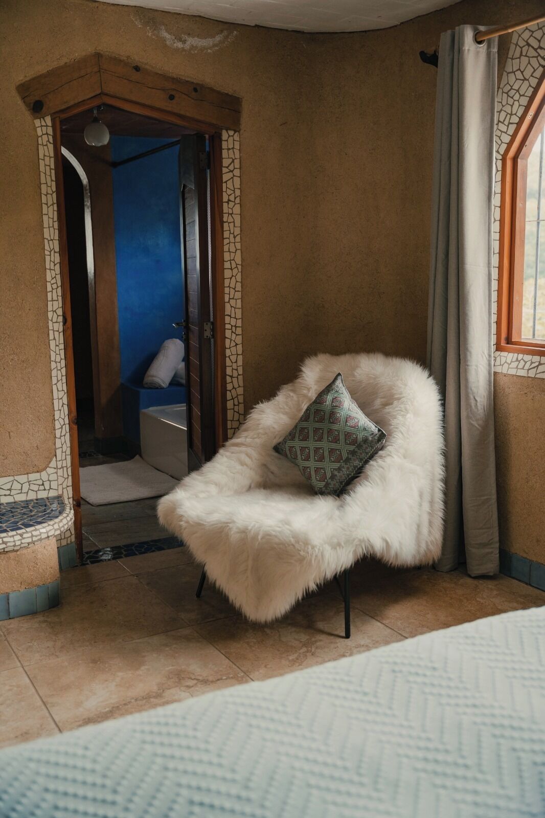 A room with a white furry chair, a patterned pillow, a window with curtains, and a view of a blue room through an archway.