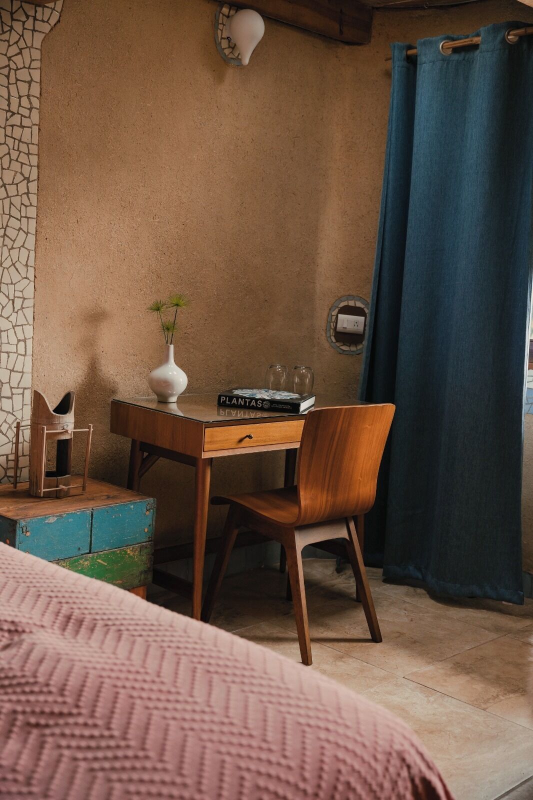 A cozy room with a wooden desk, a chair, a small white vase with plants, a blue curtain, and a stone wall.