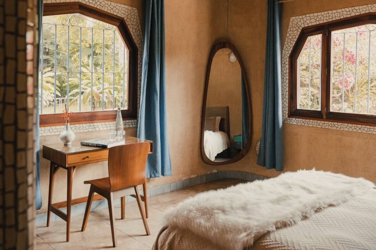 A bedroom with a wooden ceiling, a bed with a white fur throw, a desk with a chair, and a large mirror.