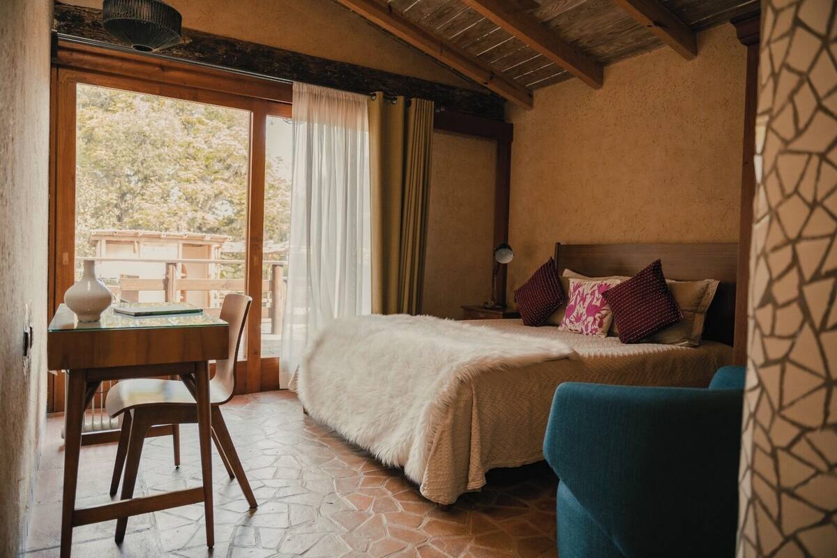 A bedroom with a bed, a desk, and a chair. There is a window with curtains, a lamp, and a view of trees outside.