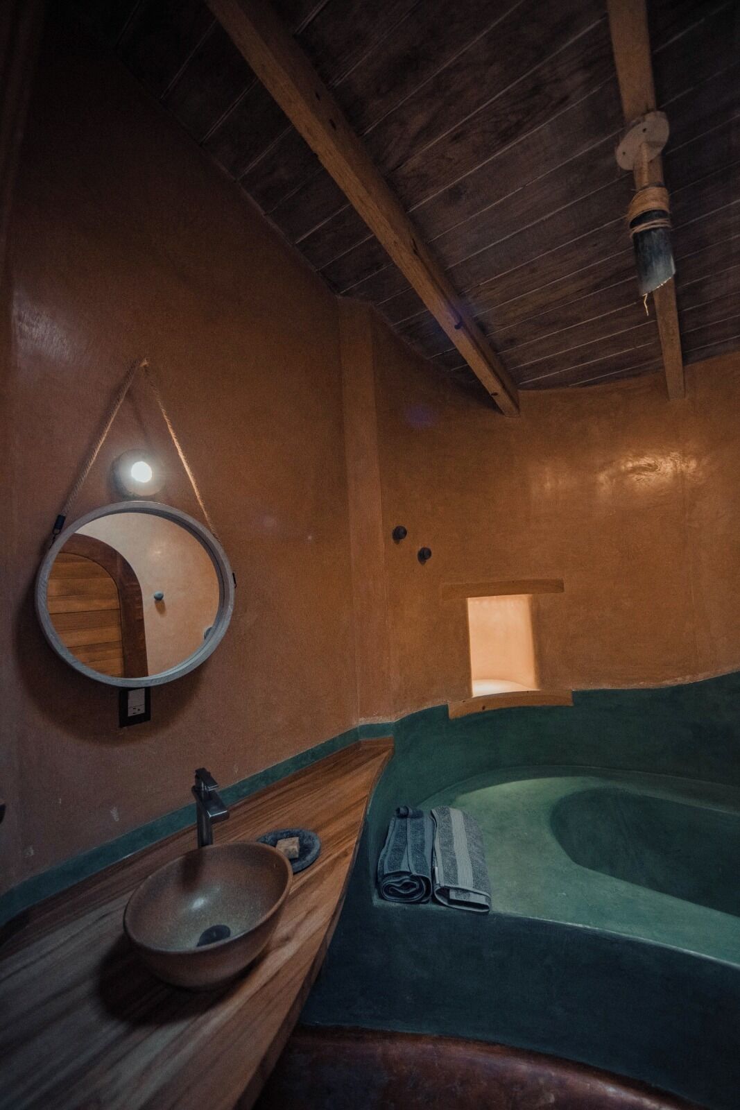 Une salle de bain rustique avec un lavabo en bois, un miroir rond, une baignoire et une petite fenêtre.