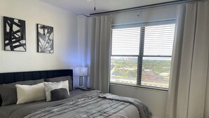 A modern bedroom with a large bed, a nightstand, a lamp, and a window with blinds.