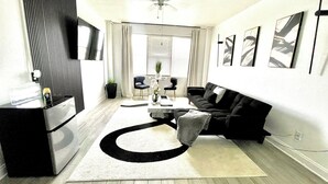 A modern living room with a black sofa, a white rug, a coffee table, and a TV on a stand.
