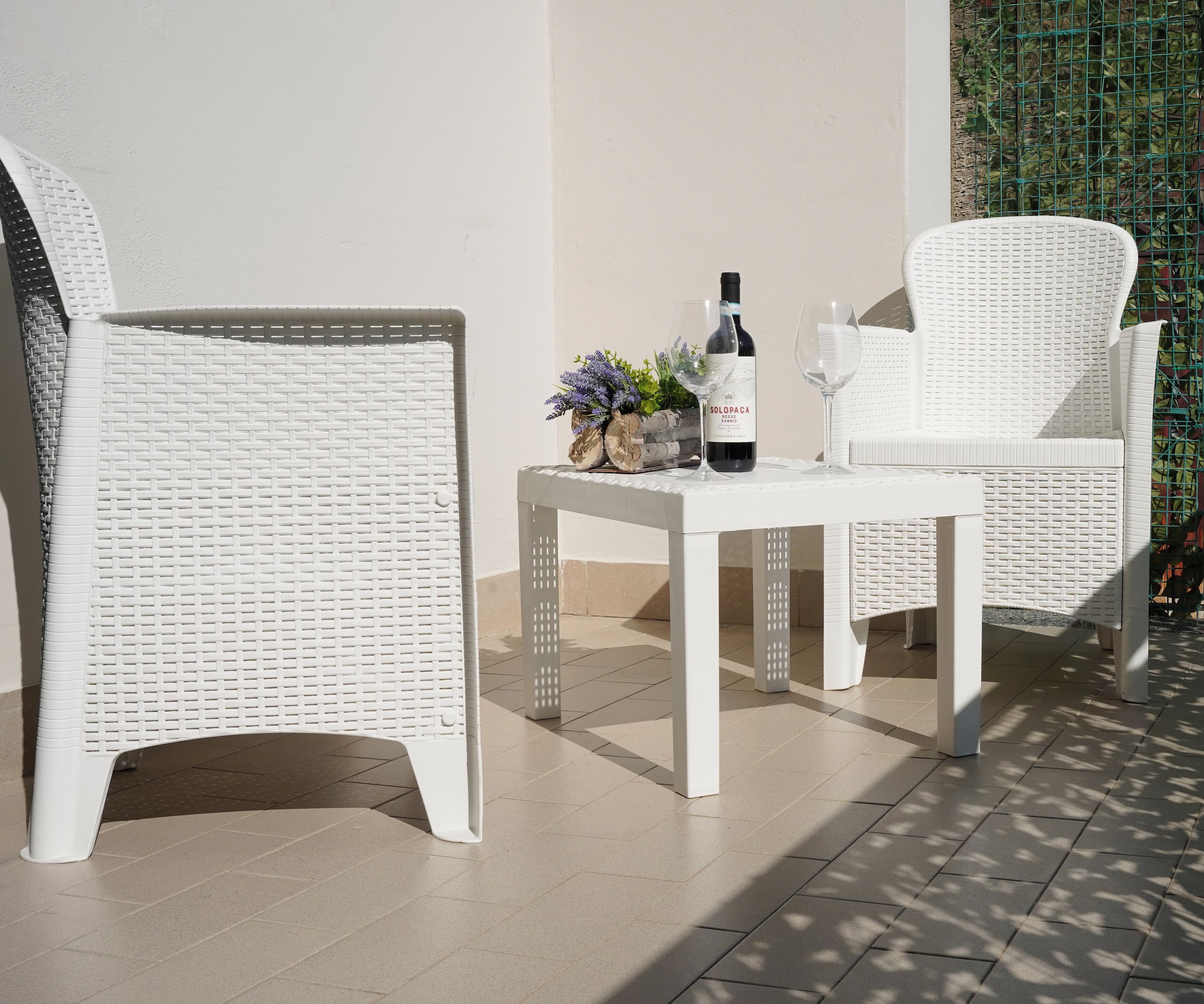 Un ensemble de meubles de jardin en osier blanc, comprenant une chaise et une table, est installé sur une terrasse carrelée, avec une bouteille de vin et un verre.