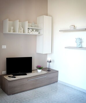 A modern living room with a flat-screen TV, a sofa, a small table with a lamp, and decorative items on shelves. - Giaro's Home (Pompei)