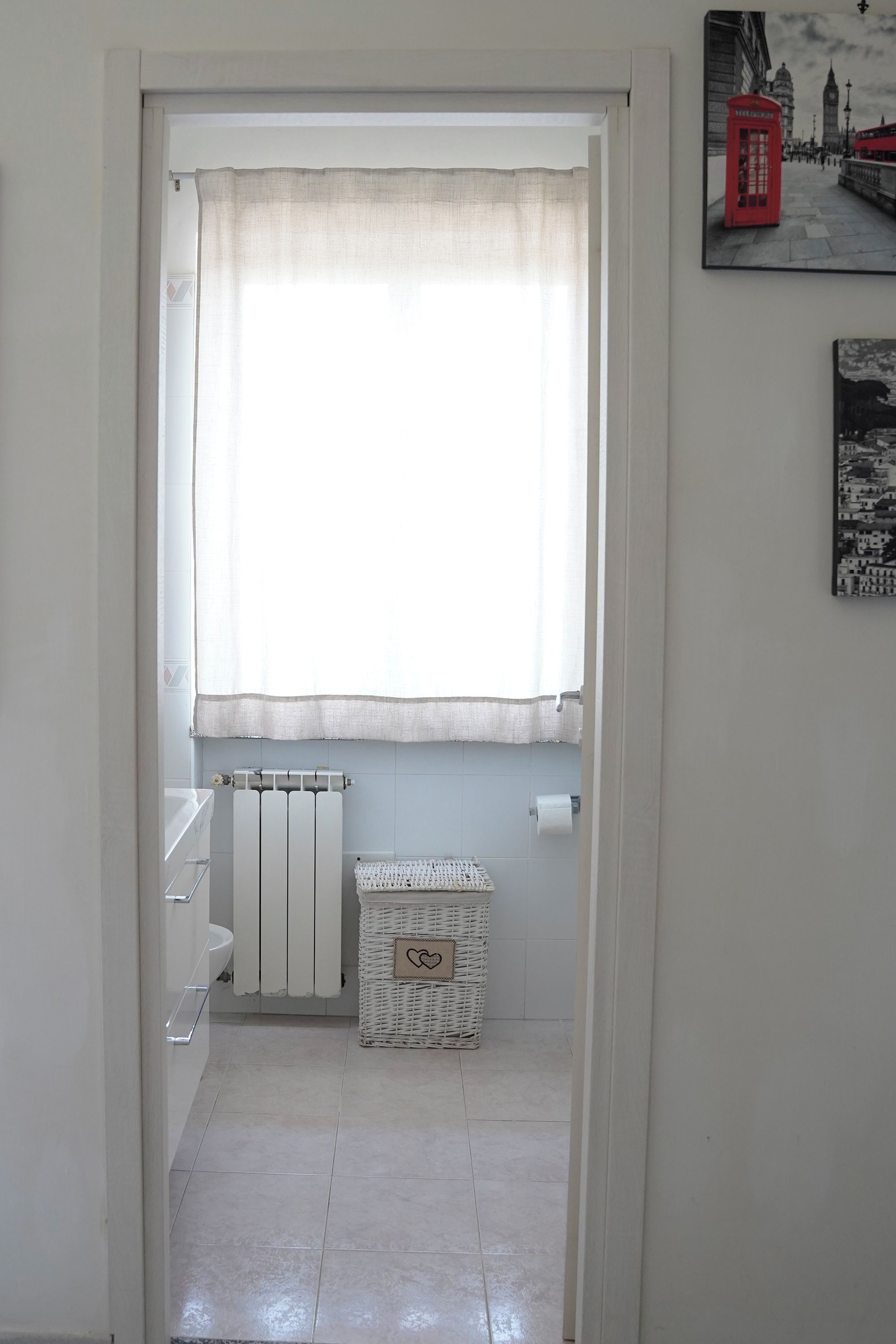 Een smalle gang met een radiator, een witte rieten mand en een raam met een dunne gordijn.