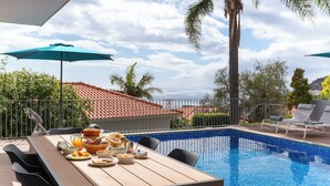 Zona pranzo a bordo piscina con tavolo apparecchiato per la colazione, con vista su una piscina e sul mare.