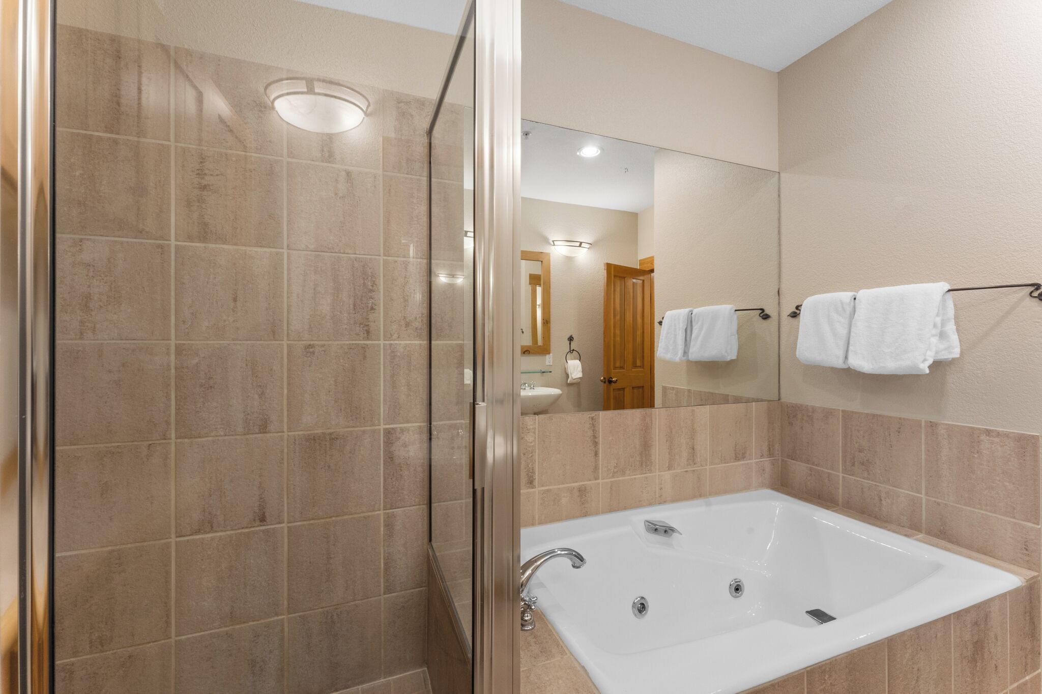 A bathroom with a glass-enclosed shower, a white bathtub, and a mirror reflecting a wooden door and towels.