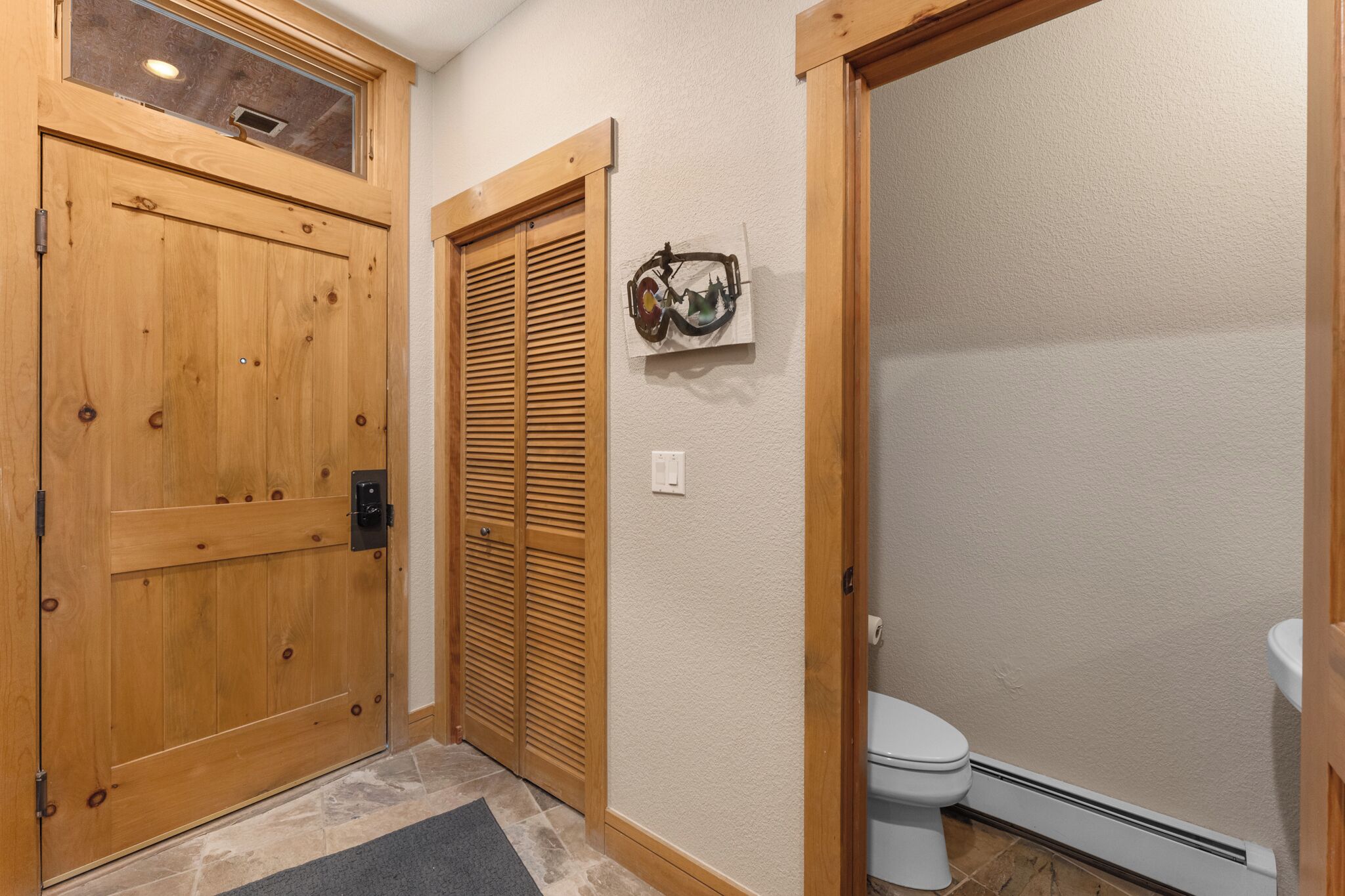 A wooden door with a window, a wooden closet with louvered doors, a white toilet, and a wall-mounted artwork.