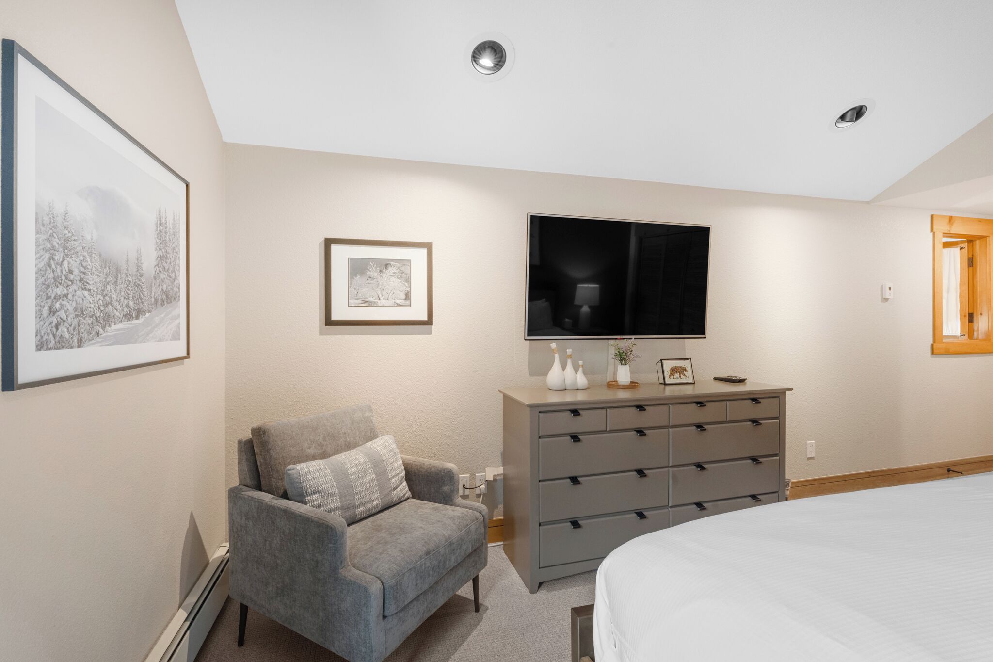 A bedroom with a bed, a gray armchair, a dresser, a flat-screen TV, and framed pictures on the wall.