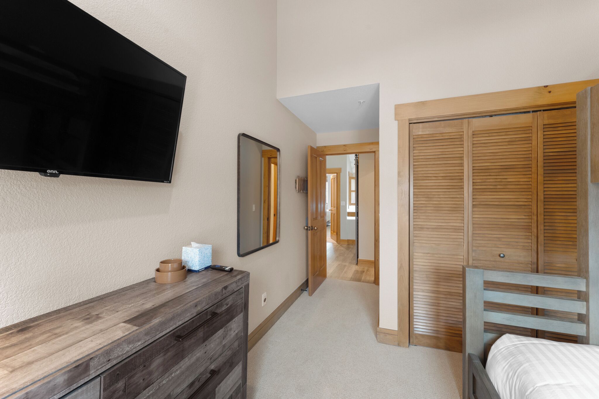 A bedroom with a bed, a wooden dresser, a TV mounted on the wall, and a wooden wardrobe.