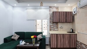 A compact living space with a green sofa, a small table with a vase of flowers, a kitchenette with wooden cabinets, and a window with a view of buildings. - Diamond Corners Serviced Apartments (Madinah)