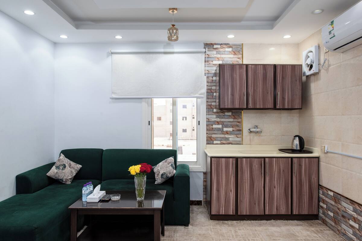 Een knus woongedeelte met een groene bank, een kleine tafel met een bloevas, een kitchenette met houten kastjes en een raam met uitzicht op gebouwen.