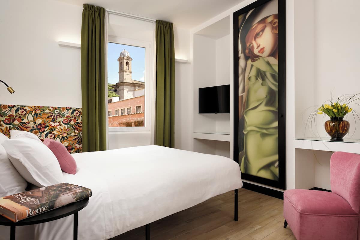 A hotel room with a bed, a TV, a painting, and a view of a building.