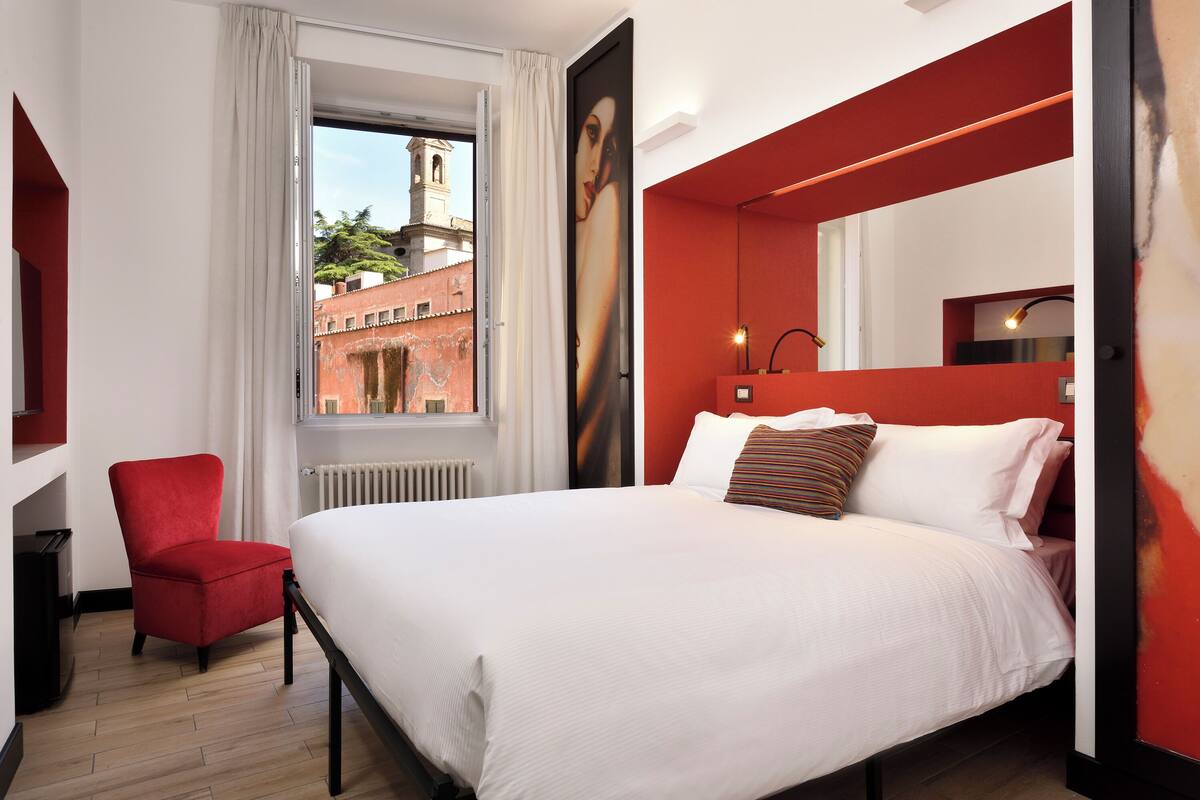 A hotel room with a large bed, a red chair, a window with a view of buildings, and a red accent wall.
