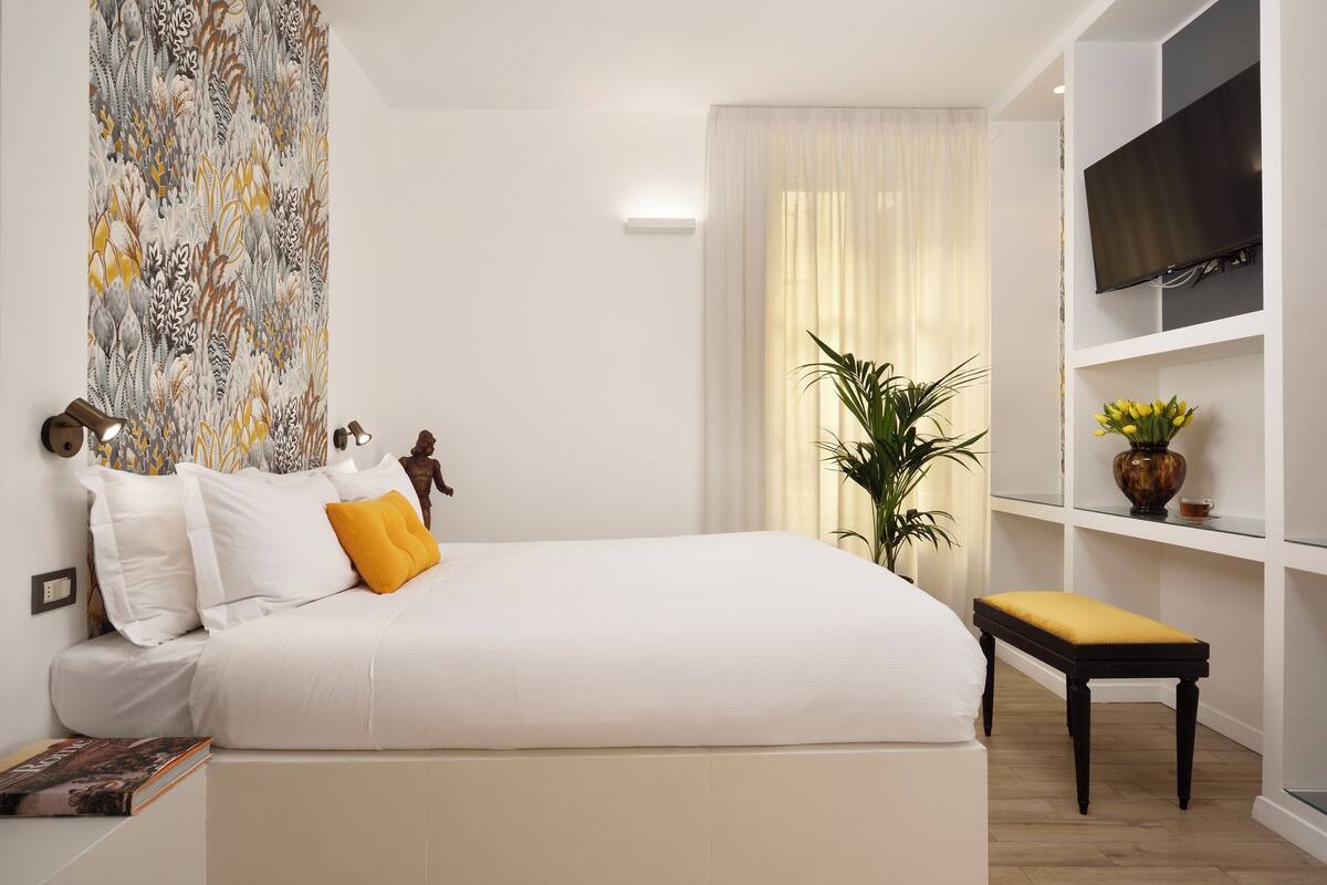 A bedroom with a bed, a bedside table, a TV mounted on the wall, a plant, and a window with curtains.