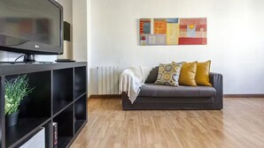 Ein modernes Wohnzimmer mit einem Flachbildfernseher auf einem schwarzen Standfuß, einem grauen Sofa mit gemusterten Kissen und einem farbenfrohen, abstrakten Gemälde an der Wand.