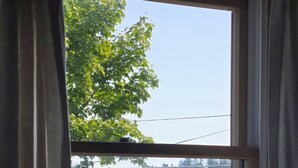Une vue à travers une fenêtre avec des rideaux, montrant un quai avec des bateaux, une voiture chargée de caisses et un bâtiment en arrière-plan.