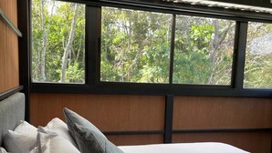 Uma cama com várias almofadas e um cobertor, uma janela com vista para árvores e uma cabeceira de madeira.