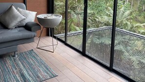 Quarto moderno com sofá cinzento, mesa redonda pequena e uma grande janela com vista para uma floresta.