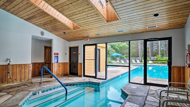 Une piscine intérieure avec un plafond et des murs en bois, une vue dégagée sur la piscine extérieure et un panneau indiquant la présence d’un sauveteur.
