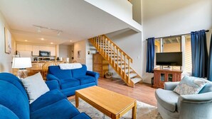 Un salon spacieux avec un canapé bleu, une table basse en bois et un fauteuil gris. Un escalier en bois mène à un niveau supérieur.