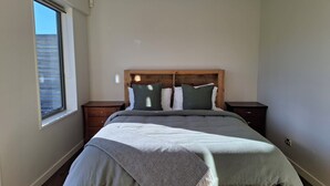Ein Schlafzimmer mit einem hölzernen Kopfteil, einem Bett mit grauer Decke, zwei Nachttischen und einem Fenster mit Jalousien.