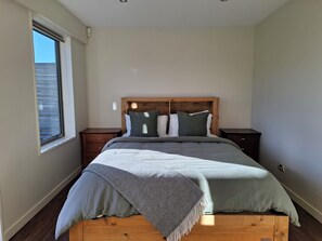Un dormitorio con cabecera de madera, cama con manta gris, dos mesitas de noche y una ventana con persianas.
