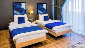 Habitación de hotel con dos camas, cada una con ropa de cama blanca y detalles azules, una mesita de noche con lámpara y obras de arte en las paredes.
