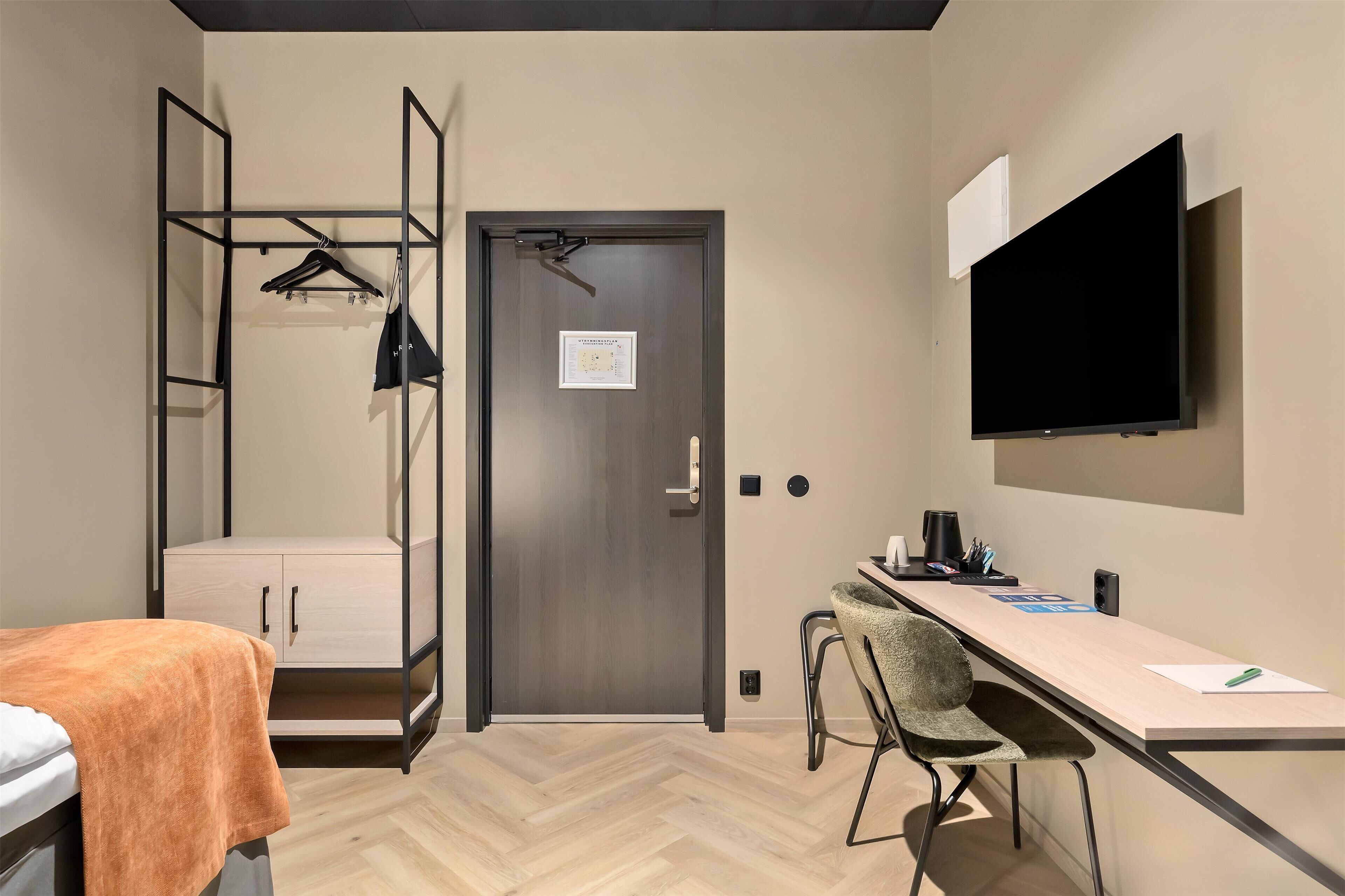 A compact hotel room with a bed, a desk, a chair, a TV, and a wardrobe with a hanging rack.