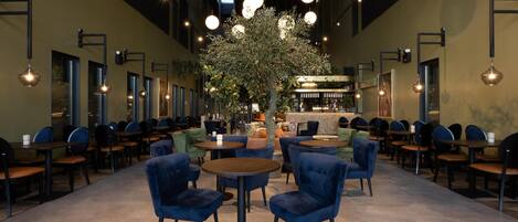 Een ruime binnenruimte met ronde tafels en stoelen, een centrale boom en een bar.