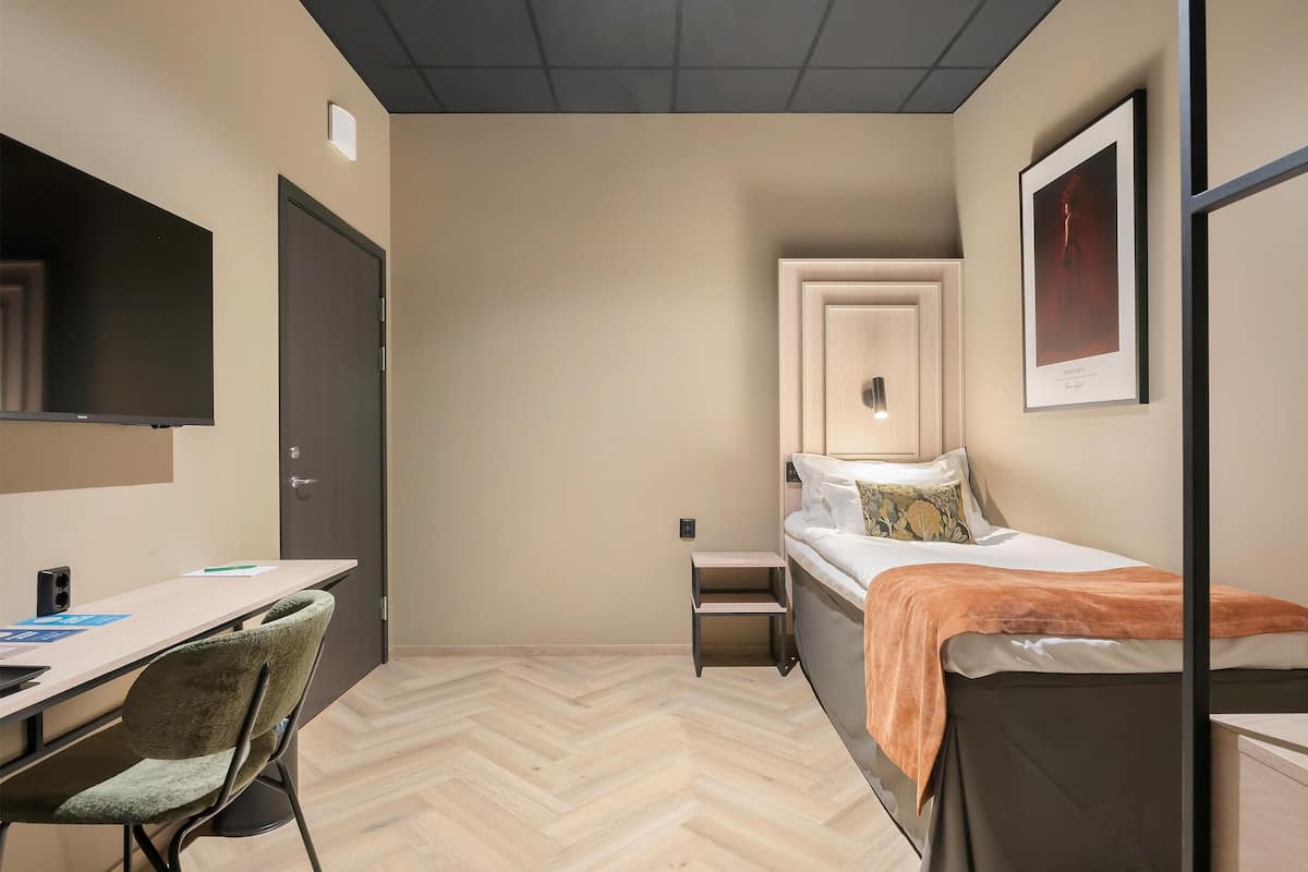 A small, modern room with a bed, a desk, and a chair. There is a door, a small table, and a framed picture on the wall.