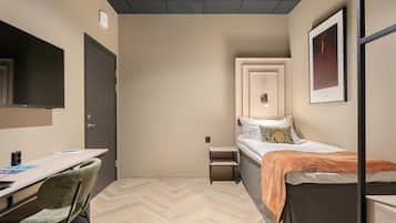Una habitación pequeña y moderna con una cama, un escritorio y una silla. Se ve una puerta, una mesita y un cuadro en la pared.