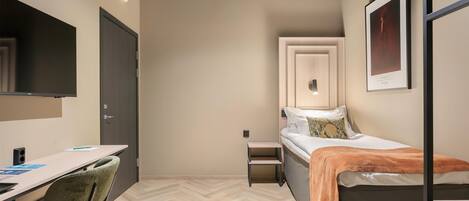 A small, modern room with a bed, a desk, and a chair. There is a door, a small table, and a framed picture on the wall.